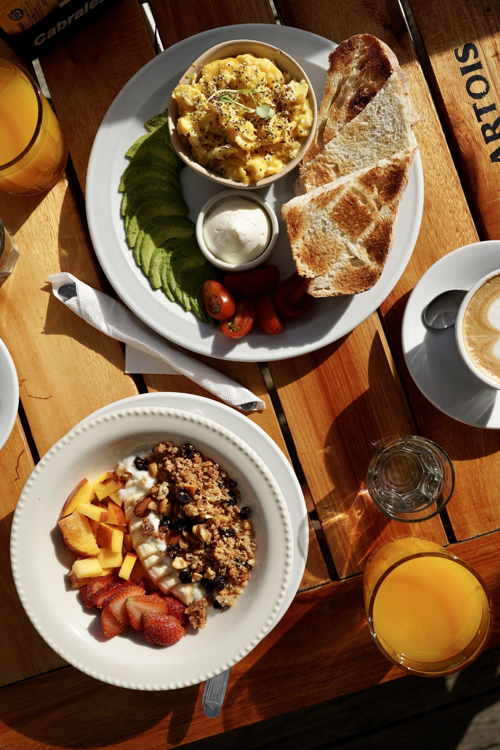 Desayuno visto desde arriba: huevos revueltos con palta y tostadas, bowl de frutas con granola, café y jugo de naranja