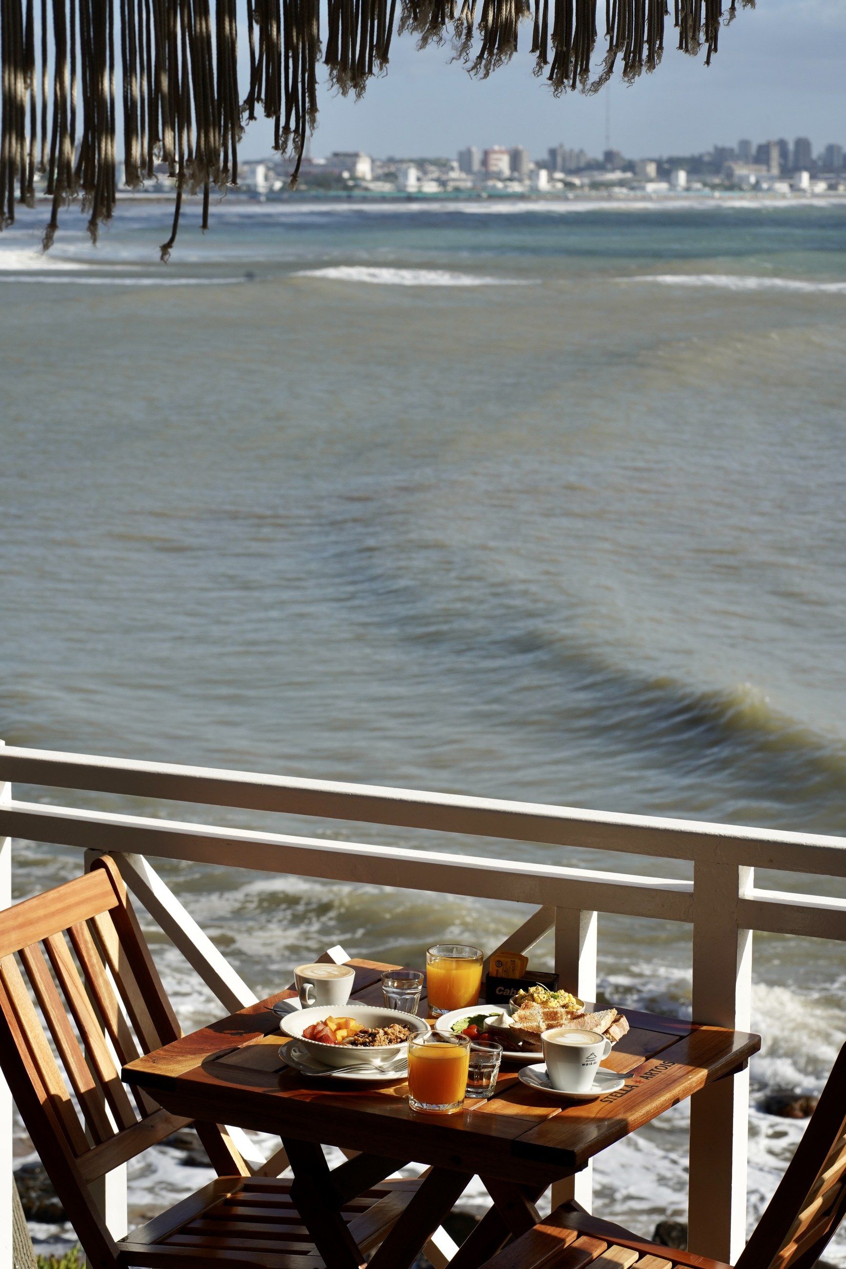 La mesa del desayuno servida en la terraza de madera, con el mar detrás de la baranda blanca