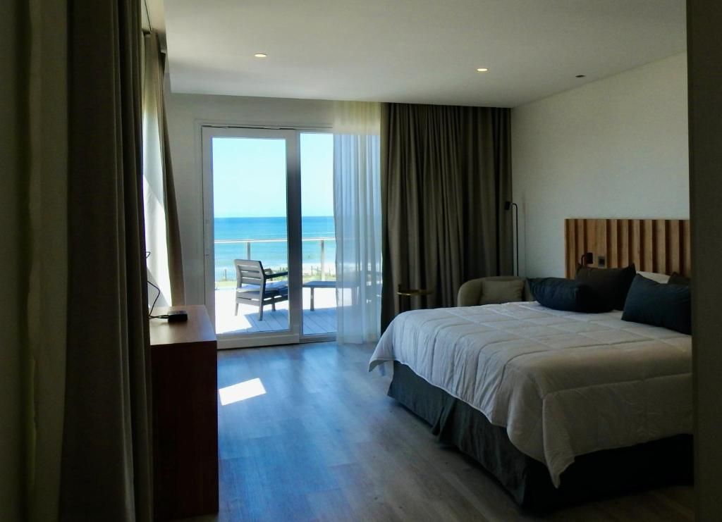 Una habitación del hotel con la cama tendida y el ventanal abierto al balcón, con el mar de fondo