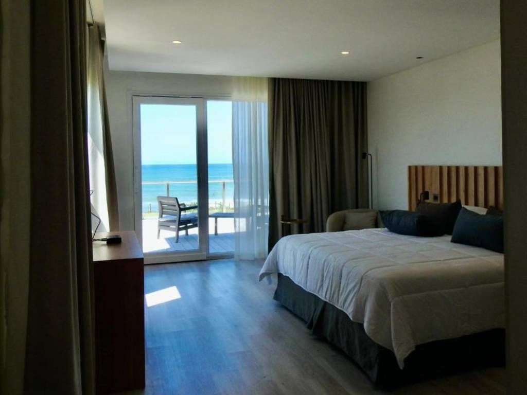 La misma habitación desde la puerta: el respaldo de madera, la cama y la salida al balcón sobre la playa